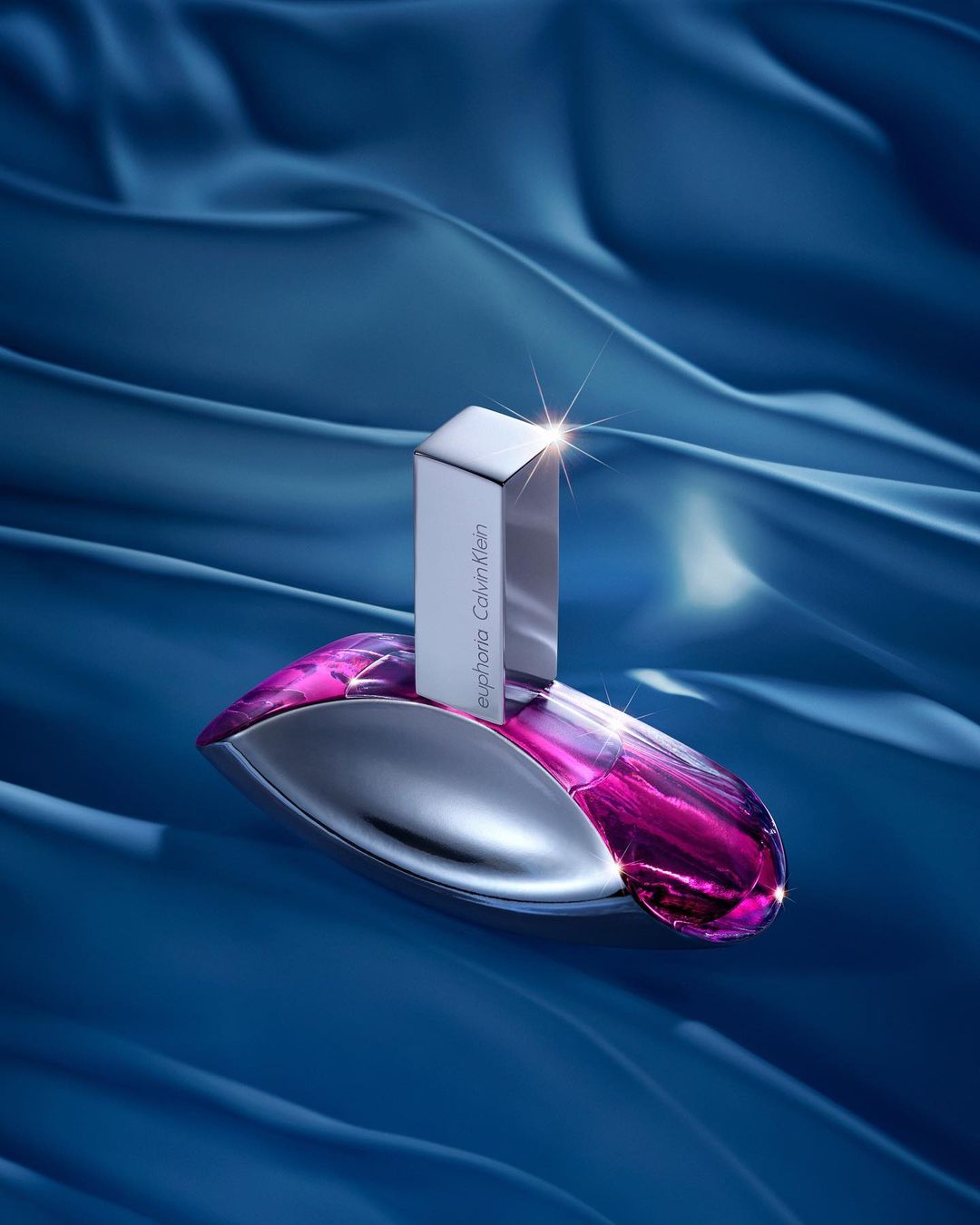 Euphoria Calvin Klein perfume bottle with a silver cap on a blue silk background in an eye-level studio shot. Beauty photography.