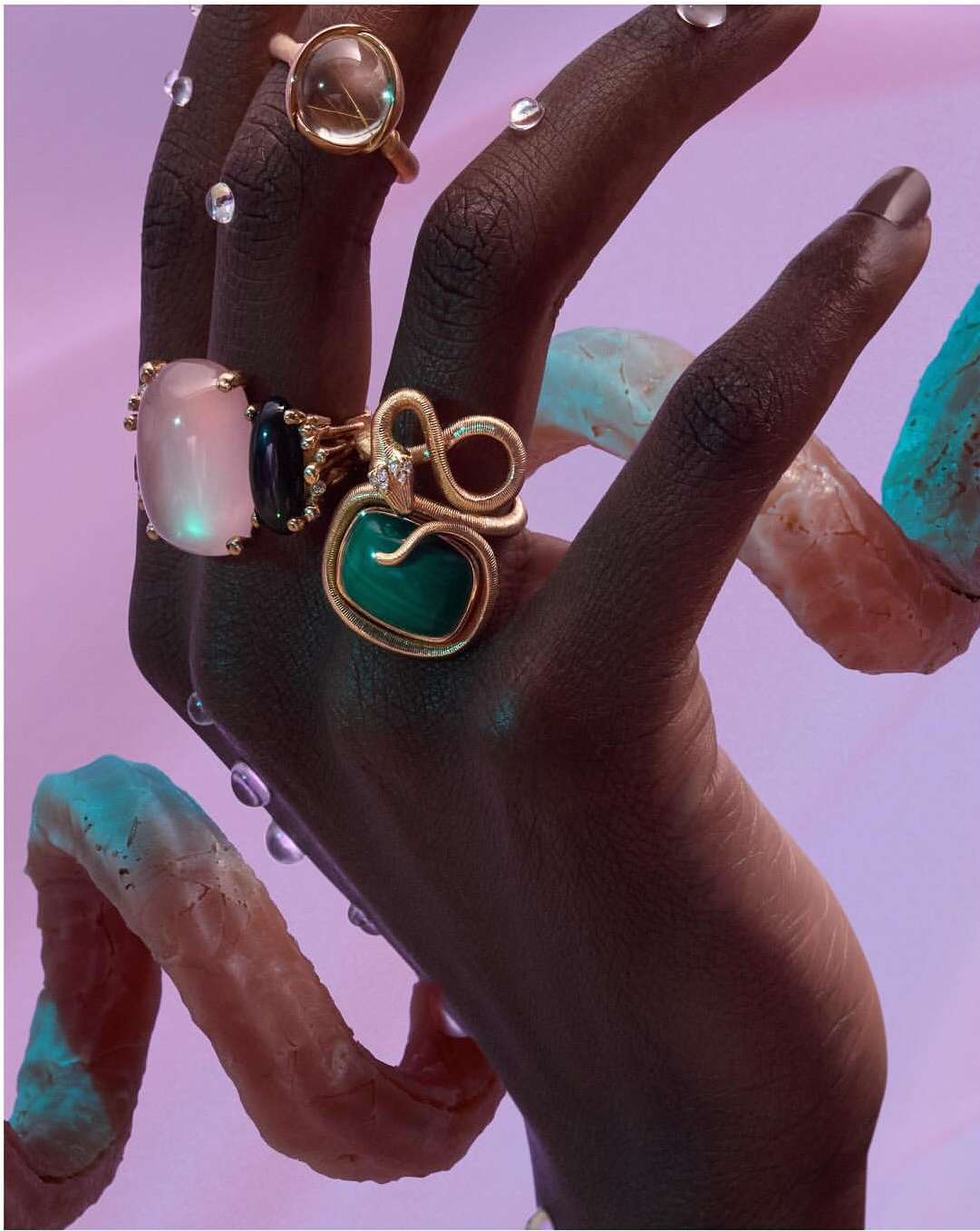 An upward view of a dark-skinned hand adorned with gemstone rings in gold settings and small dew drops, against a lilac backdrop.