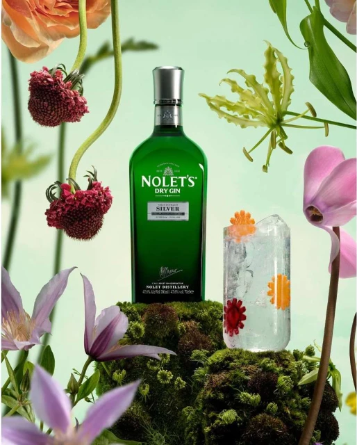 A bottle of Nolet's Dry Gin next to a gin cocktail with ice, and floral arrangements, set on mossy ground, in a studio.