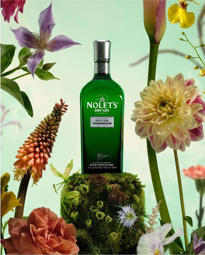 Still life of a green glass gin bottle on a mossy mound, surrounded by colorful flowers against a light green background.