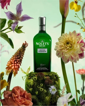 Still life of a green glass gin bottle on a mossy mound, surrounded by colorful flowers against a light green background.