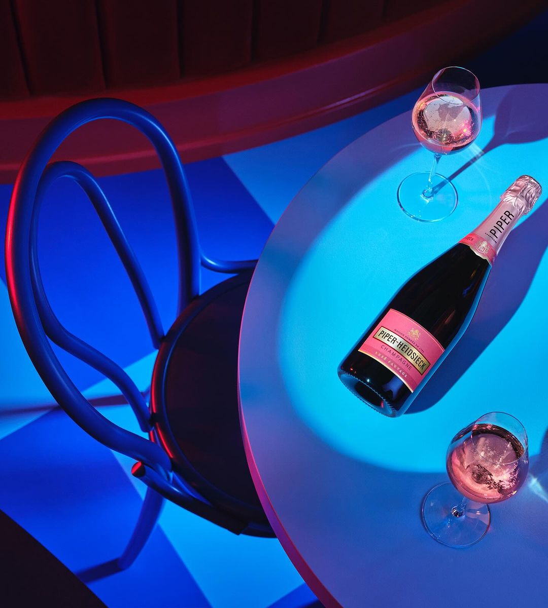Overhead shot of Piper Heidsieck Champagne bottle and two glasses on a round table with blue and pink lighting and a blue chair.