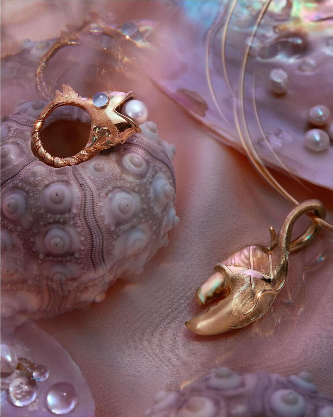 Still life of gold jewelry: a fish ring with a pearl, and a crab claw pendant necklace, displayed on a sea urchin with a pink background.