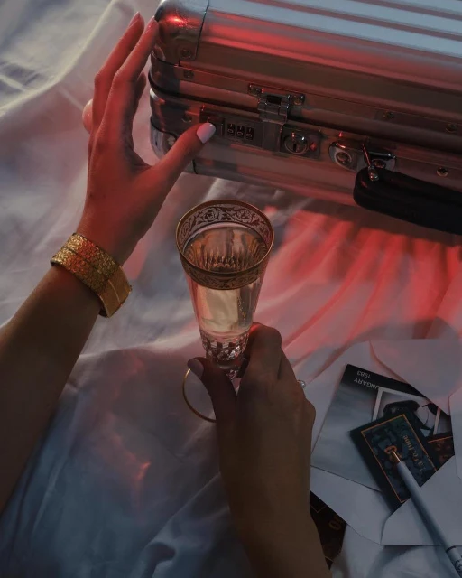 Close-up shot of adult hands holding a champagne flute next to an aluminum briefcase, cards, and a pen on a white sheet.