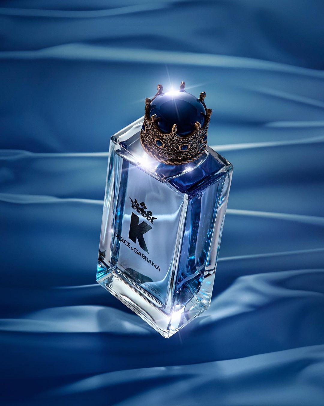 Dolce & Gabbana K eau de parfum bottle on blue satin. The men's fragrance features a decorative crown cap and a geometric glass bottle.