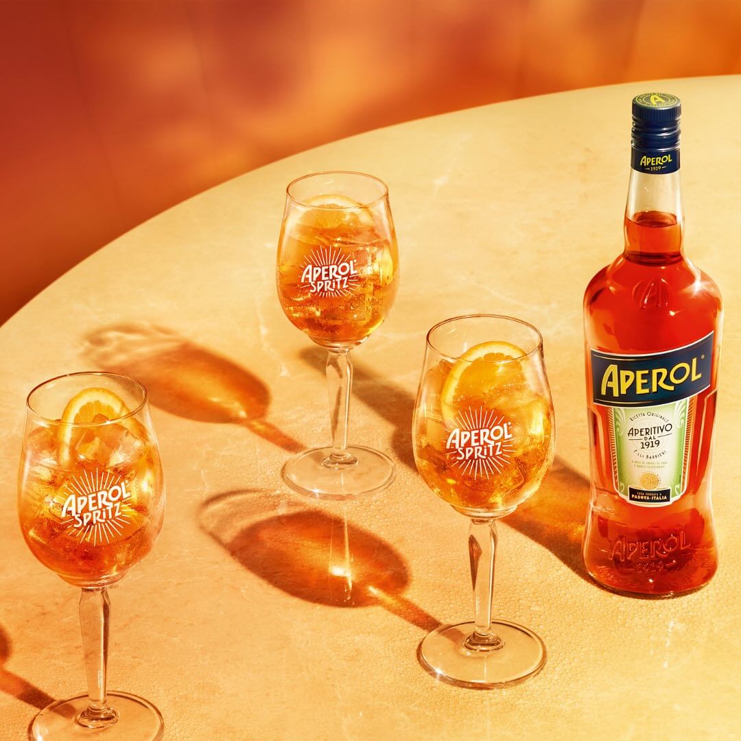 Three Aperol Spritz cocktails with orange slices and a bottle of Aperol on a tan surface, bright, commercial food and beverage shot.