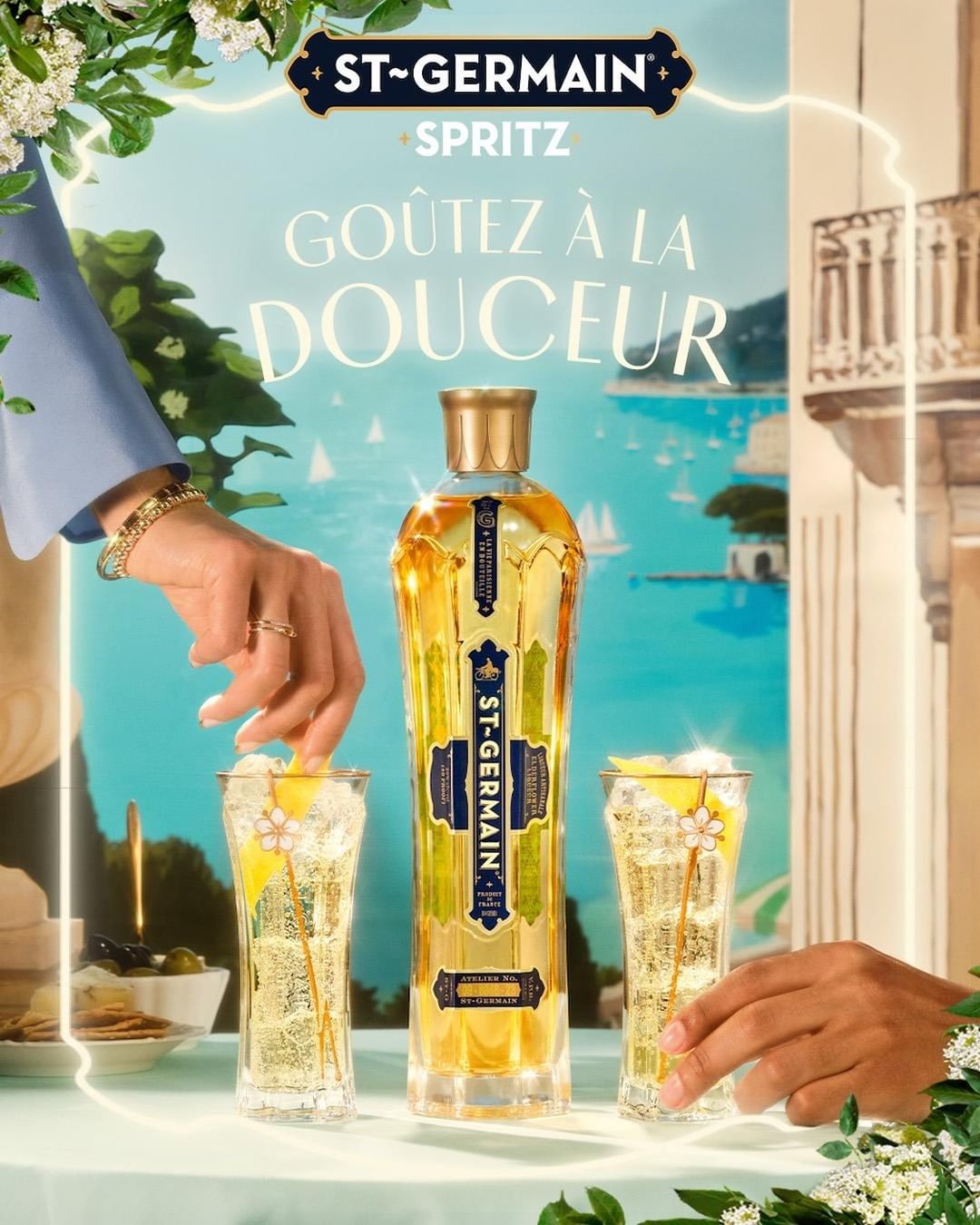 St-Germain Spritz ad featuring a bottle, two cocktails, and hands placing lemon slices in glasses against a coastal vista backdrop.