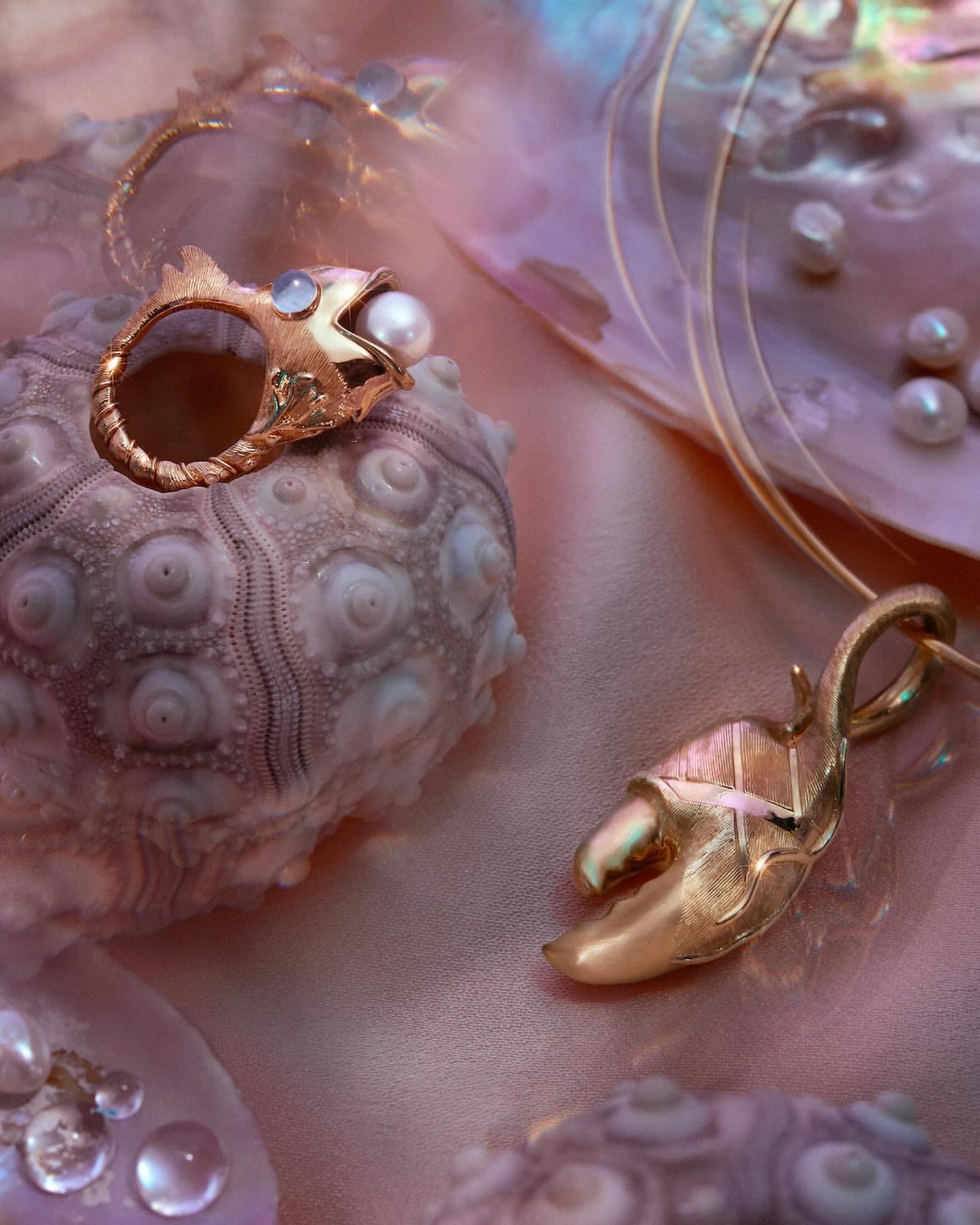 Close-up of a gold ring with a pearl, a gold crab pendant, and sea shells on a pink satin background. Jewelry product shot.