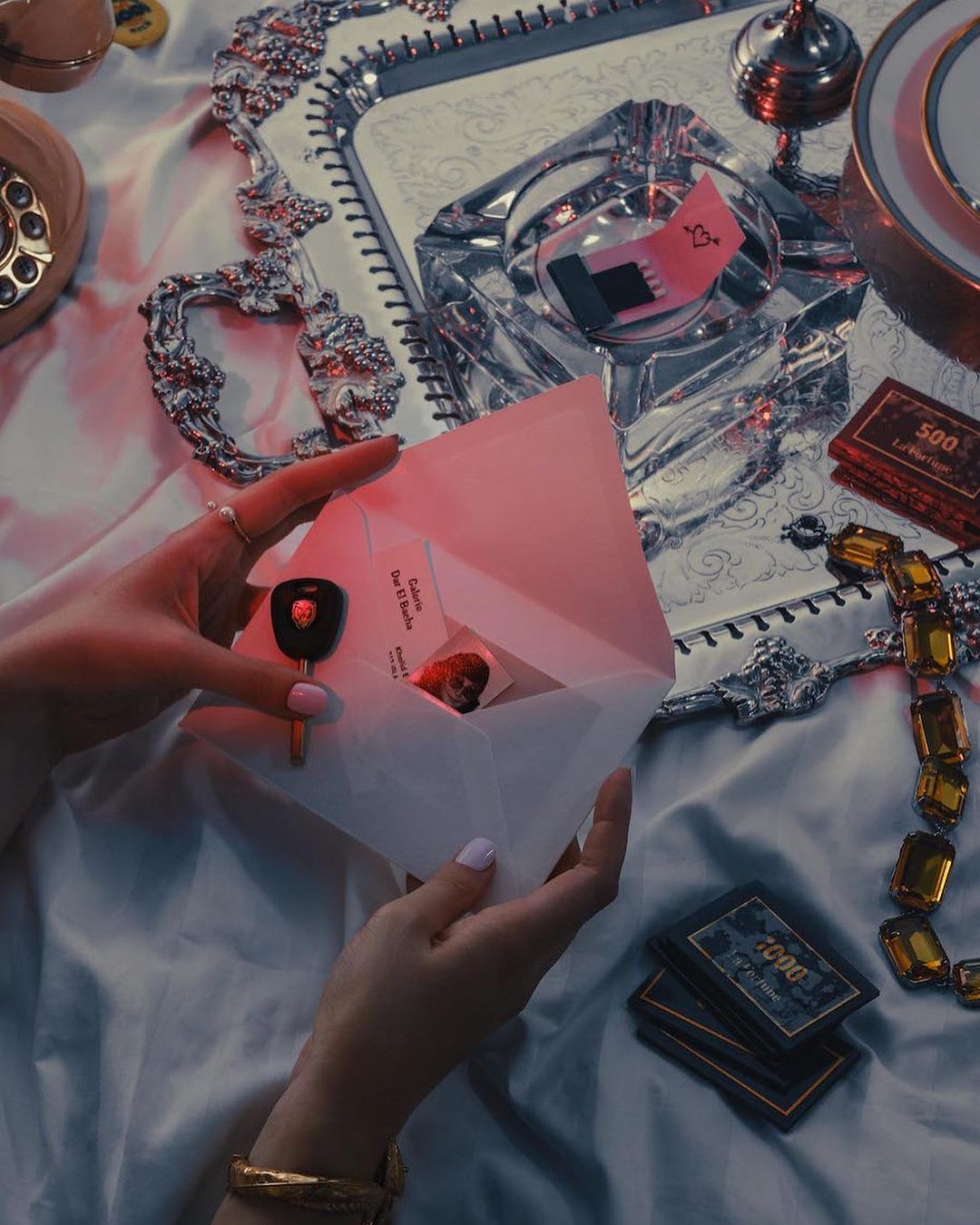 Overhead shot of hands opening an envelope with a car key and cards on a silky background, accessorized with jewelry and a vintage phone.
