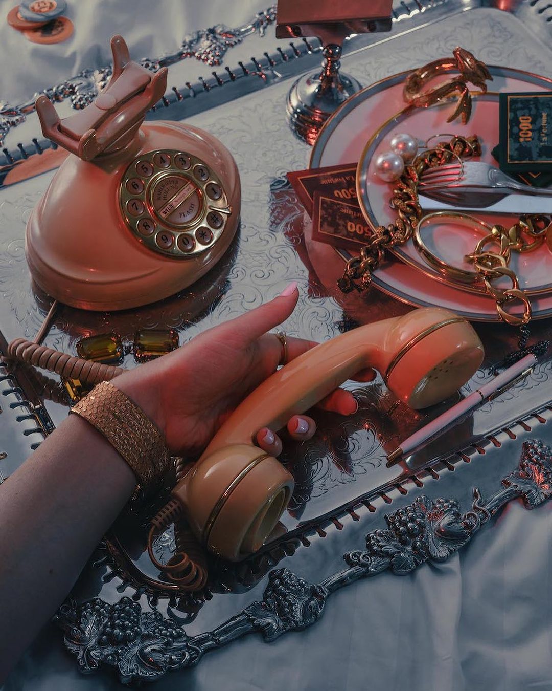 A vintage peach-colored rotary telephone is held by a hand with gold jewelry, set on a silver tray with retro objects, creating a nostalgic aesthetic.