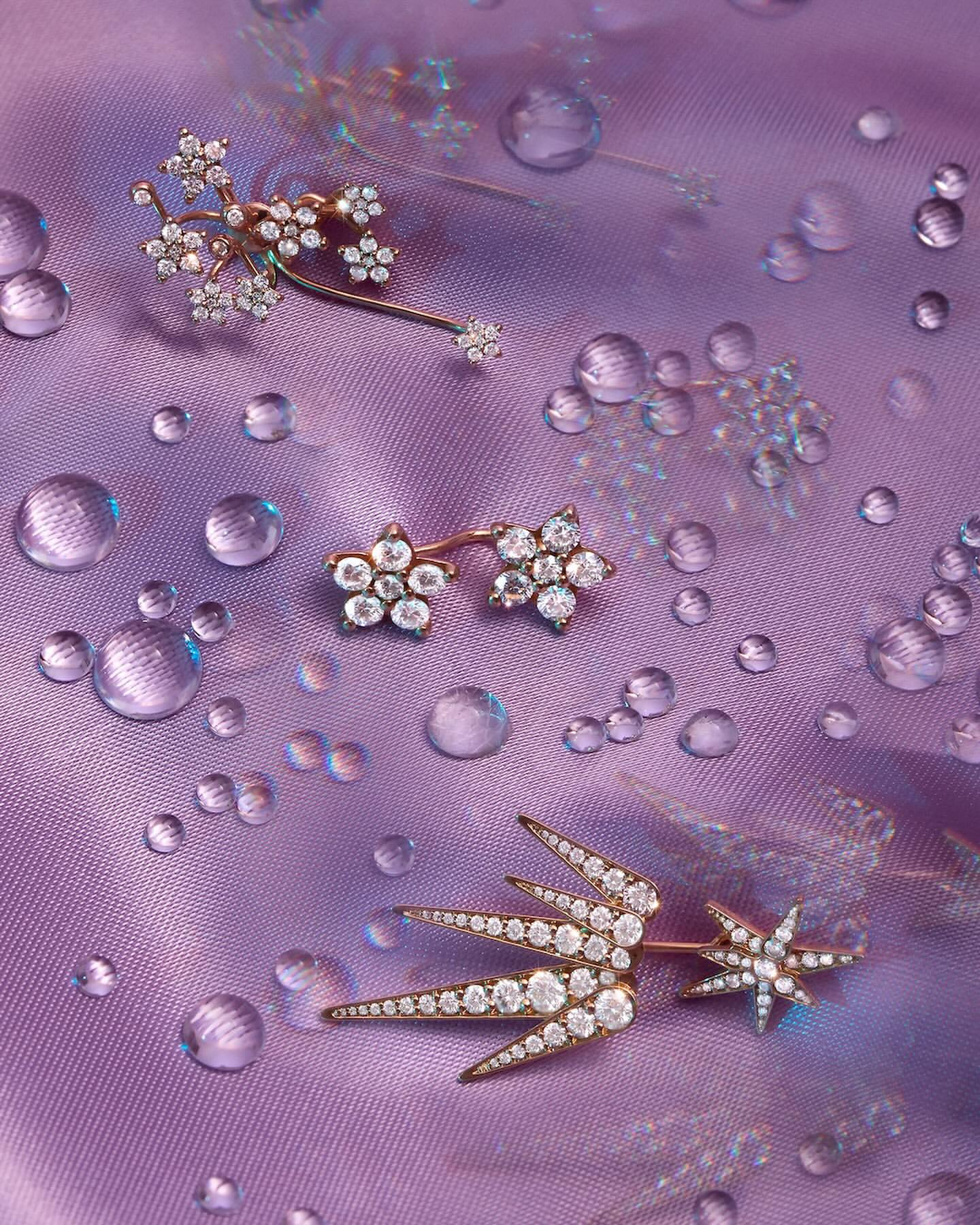 Close-up of three floral-inspired gold-toned and diamond-encrusted earrings of different sizes, scattered on a light purple silk fabric with water droplets.