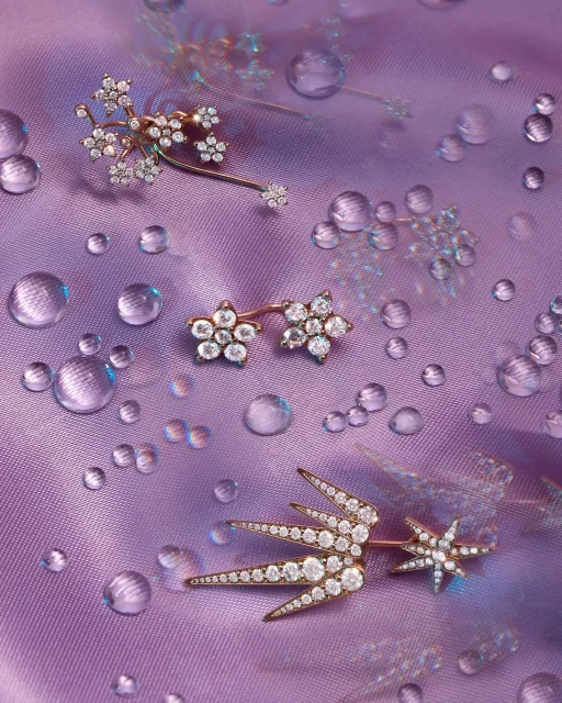 Close-up of three floral-inspired gold-toned and diamond-encrusted earrings of different sizes, scattered on a light purple silk fabric with water droplets.