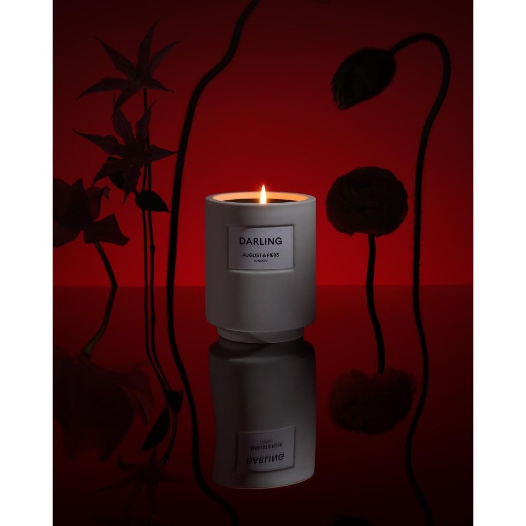 A grey candle sits on a reflective surface with floral accents against a red backdrop, offering a moody, artistic feel.