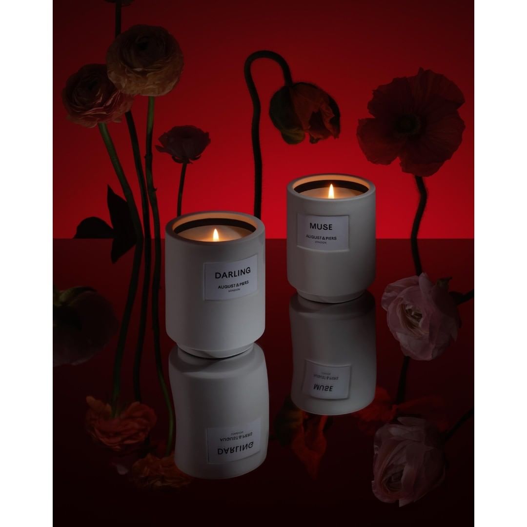 Two lit candles sit on a reflective surface surrounded by colorful flowers and a red background, creating an elegant and moody scene.