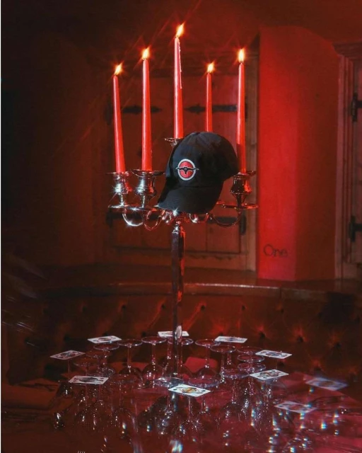 Still life photo showcasing a black baseball cap atop a silver candelabra with lit red candles, surrounded by playing cards and glassware.