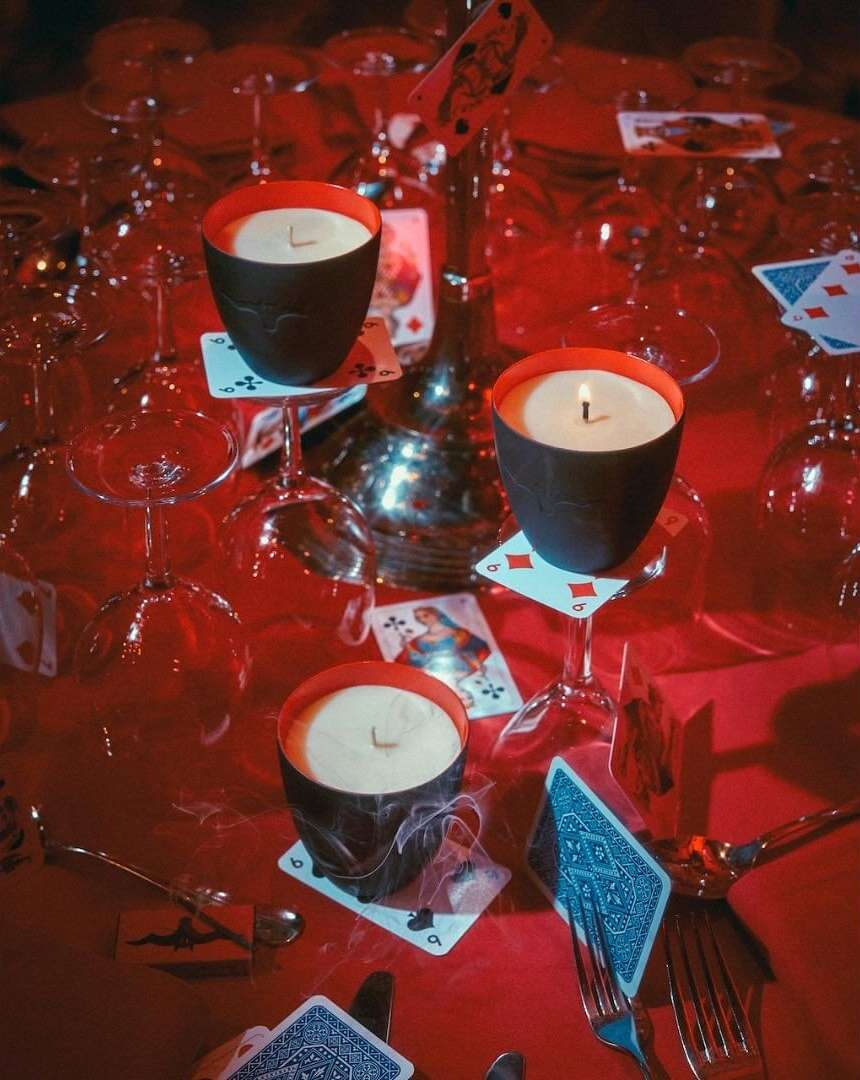 Table setting with playing cards, wine glasses, silverware, lit candles in black holders with red interiors, on red tablecloth.