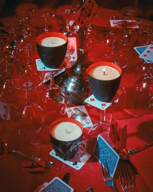 Table setting with playing cards, wine glasses, silverware, lit candles in black holders with red interiors, on red tablecloth.