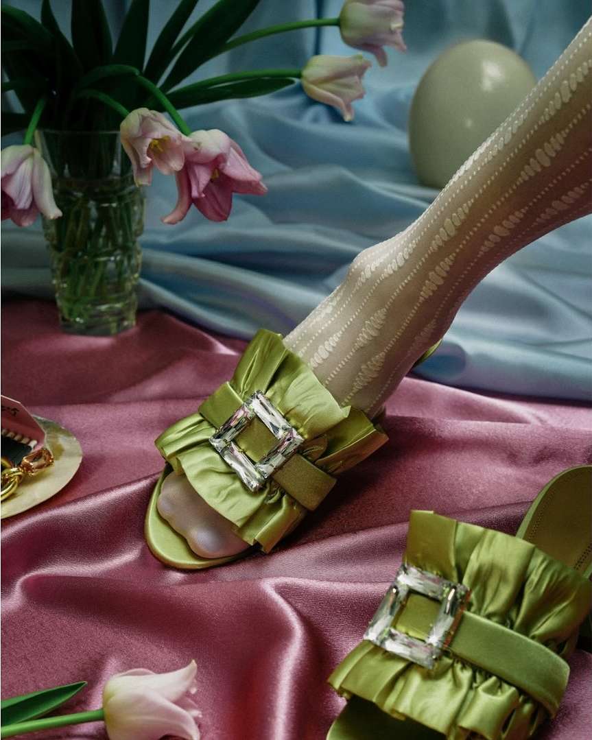 Still life of green satin ruffled mules with crystal buckles, patterned stockings, tulips in a vase, and a white sphere.