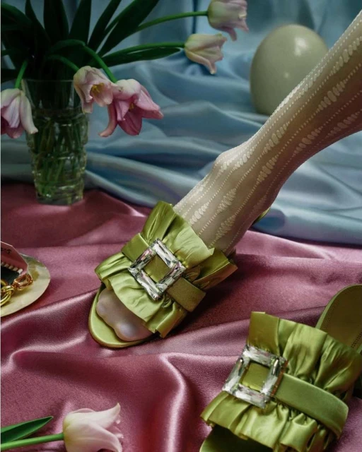 Still life of green satin ruffled mules with crystal buckles, patterned stockings, tulips in a vase, and a white sphere.