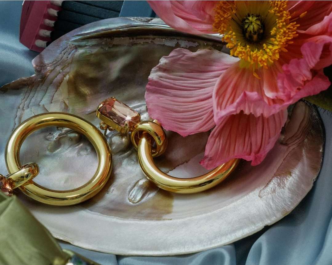 Close-up of gold hoop earrings with gemstone accents, set on a seashell next to a pink flower and light blue satin fabric.