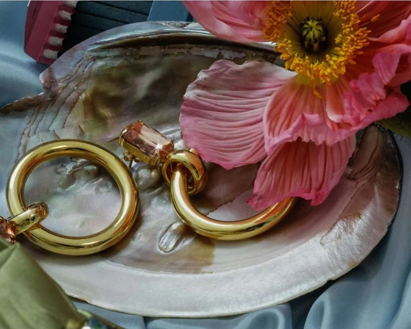 Close-up of gold hoop earrings with gemstone accents, set on a seashell next to a pink flower and light blue satin fabric.