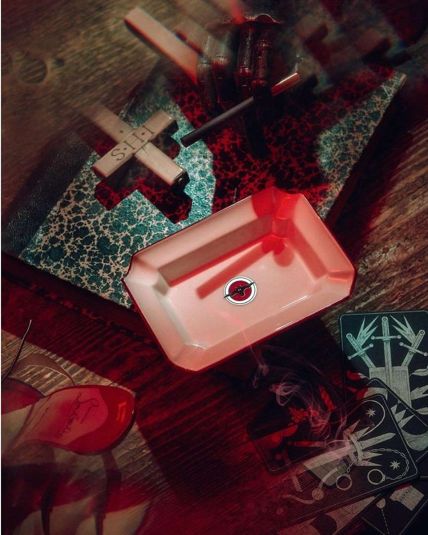 Overhead shot of an arrangement featuring a white ashtray with red design, tarot cards, and a cross on a wood surface.