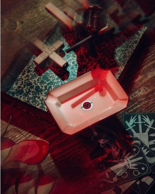 Overhead shot of an arrangement featuring a white ashtray with red design, tarot cards, and a cross on a wood surface.