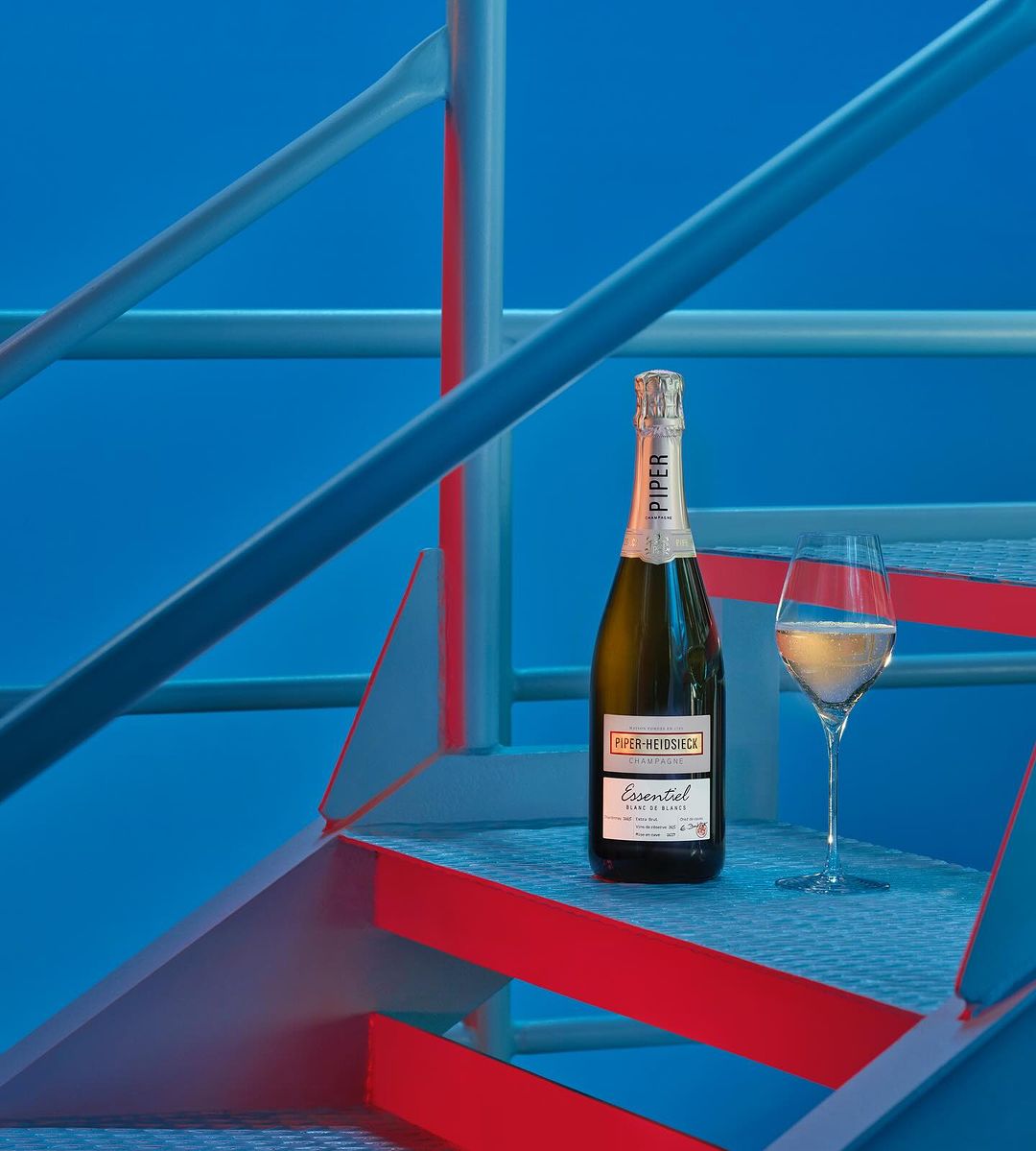 Bottle of Piper Heidsieck champagne with a champagne flute on steps, bathed in blue and red lighting, creating a modern still life.