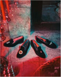 Four black velvet loafers with embroidered motifs arranged on a stone hearth under dramatic red and teal lighting.