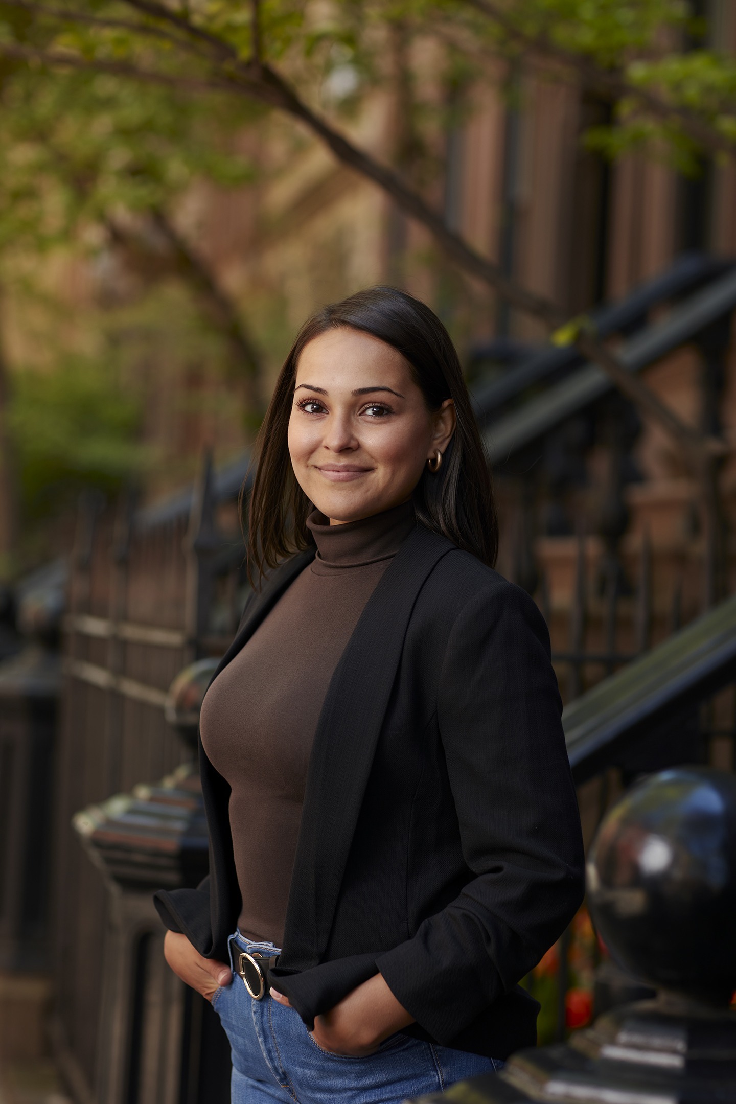 A woman in a brown turtleneck and black blazer stands smiling outside with dark hair and blue jeans in an urban environment.