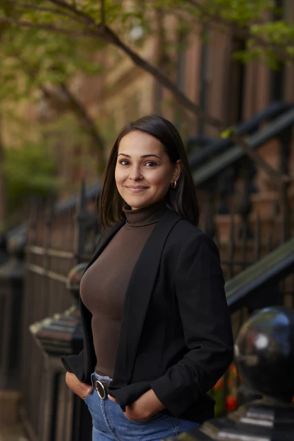 A woman in a brown turtleneck and black blazer stands smiling outside with dark hair and blue jeans in an urban environment.