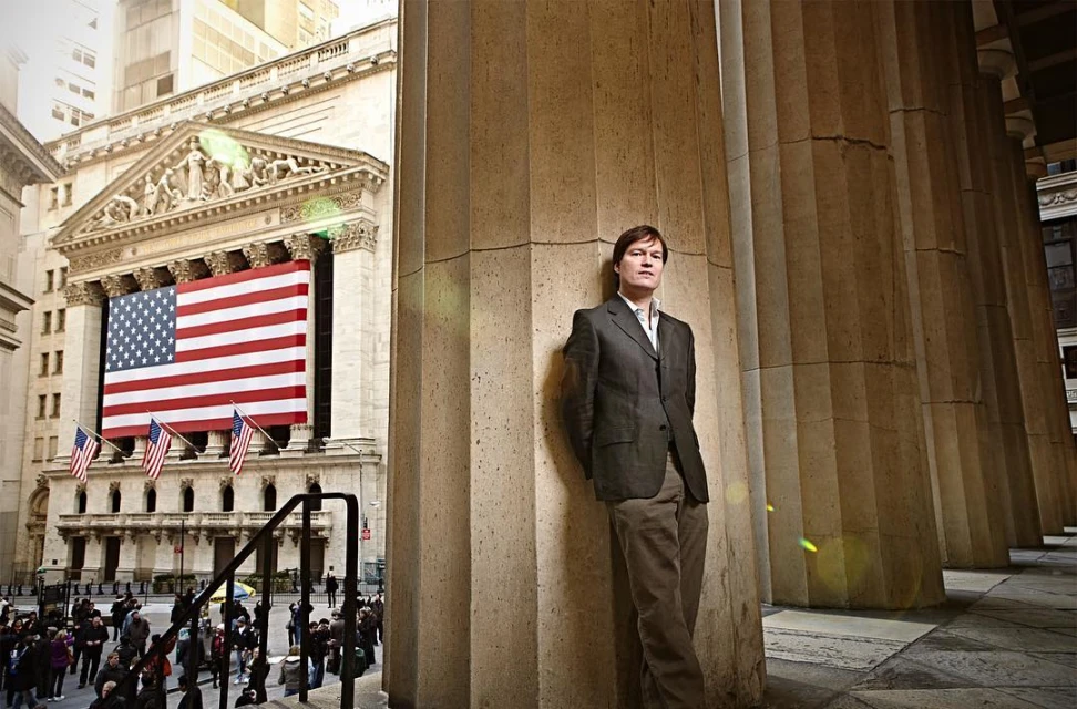 Man in suit leans against a column outside the New York Stock Exchange. An American flag adorns the building in the background.