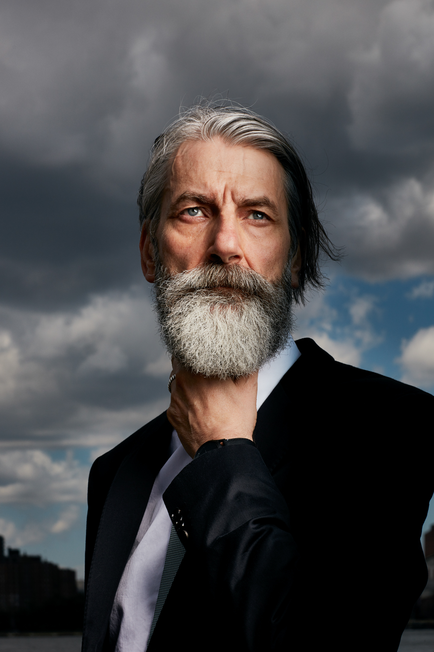 Dramatic portrait of a mature man with grey hair and beard in a black suit, posing outdoors against a cloudy sky and waterfront.