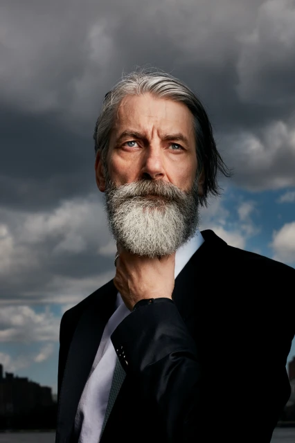 Dramatic portrait of a mature man with grey hair and beard in a black suit, posing outdoors against a cloudy sky and waterfront.
