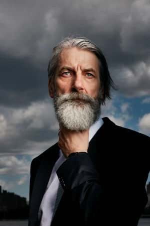 Dramatic portrait of a mature man with grey hair and beard in a black suit, posing outdoors against a cloudy sky and waterfront.