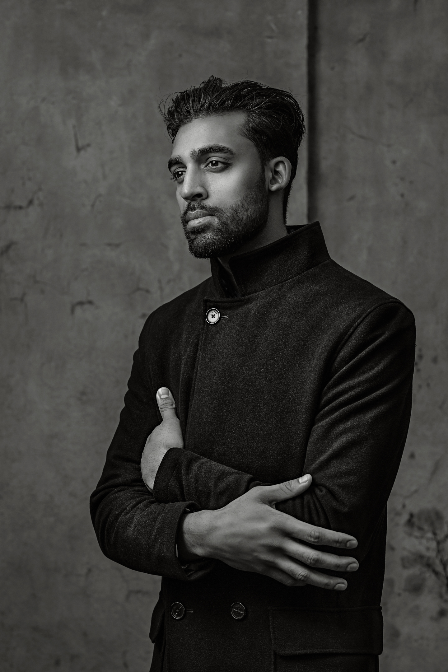 Monochrome portrait of a South Asian man in a dark high collar coat with arms crossed against a textured concrete wall.