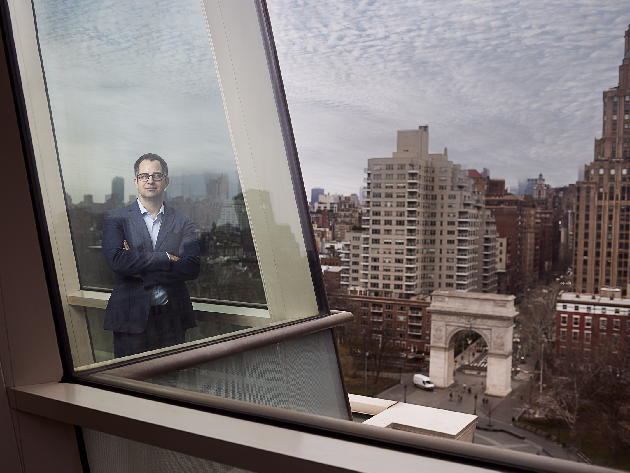 Man in a navy blue suit and glasses standing inside by a large angled window overlooking a city street with an arch monument.