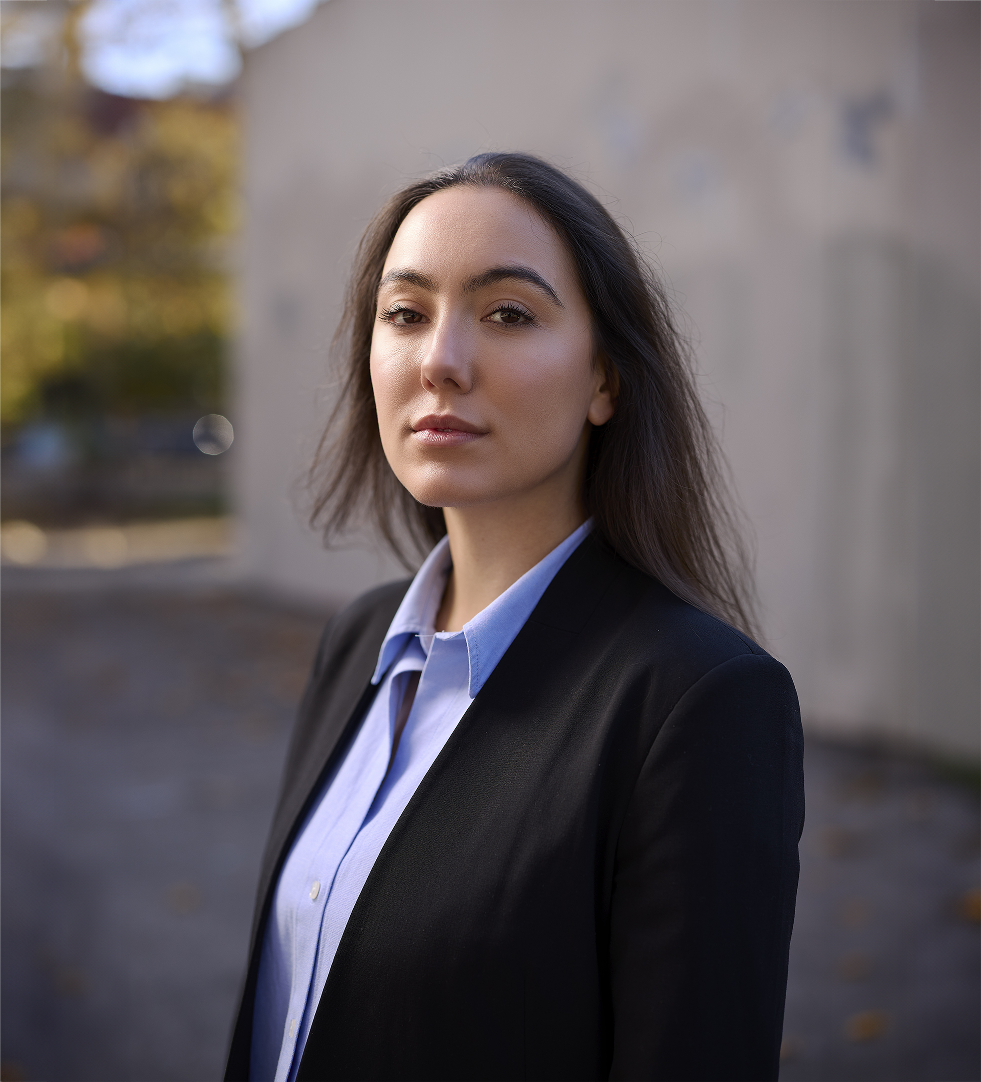 Portrait of a young woman with dark hair wearing a black blazer and light blue collared shirt, standing outdoors in a blurred background.