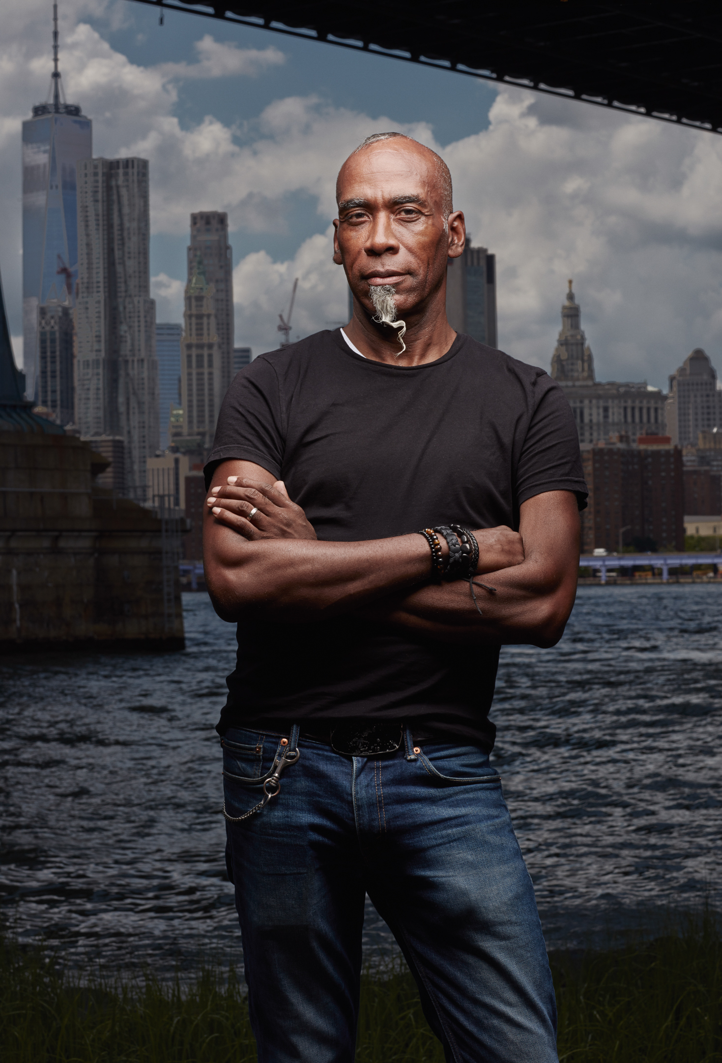 An adult Black man stands with arms crossed in jeans and a black shirt, posing outdoors with a river and cityscape backdrop.