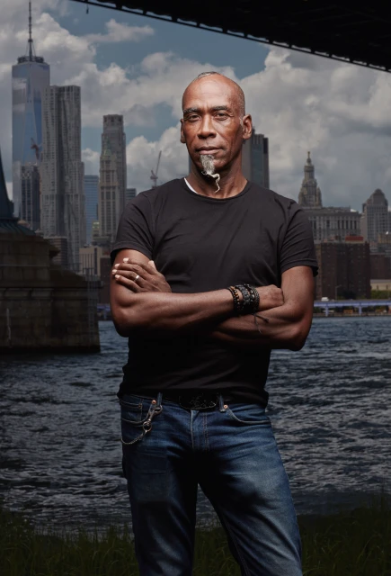 An adult Black man stands with arms crossed in jeans and a black shirt, posing outdoors with a river and cityscape backdrop.