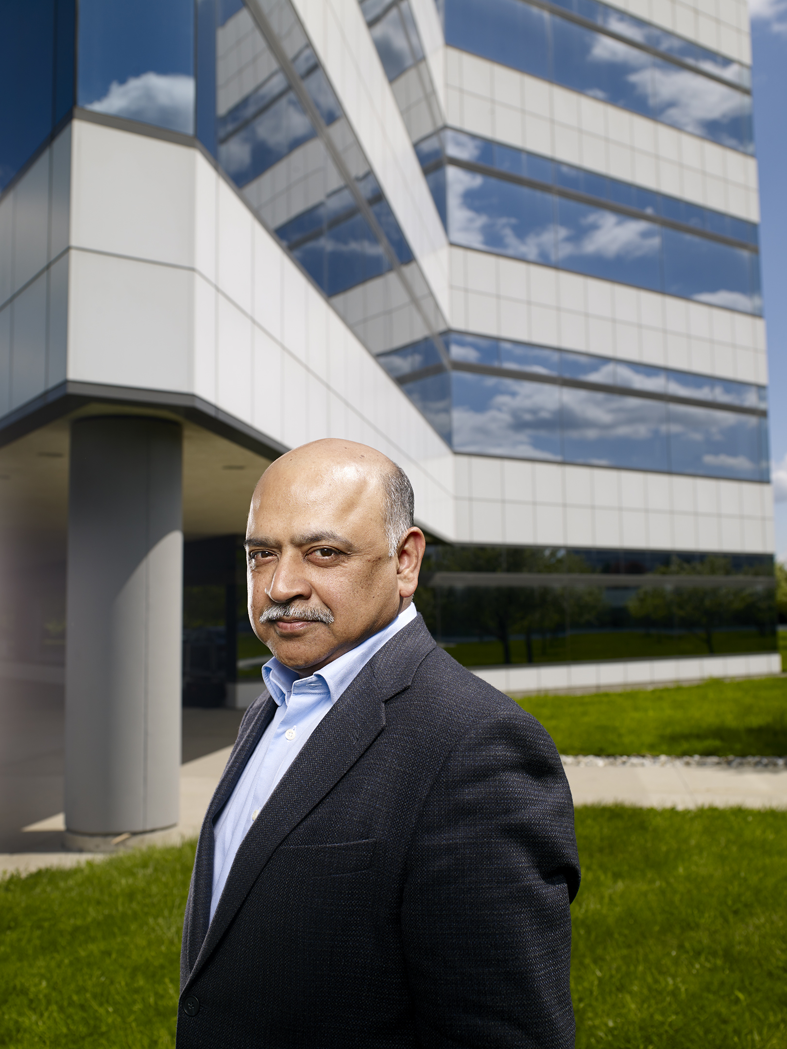 Portrait of a middle-aged man in a suit and button-down shirt standing in front of an office building and a green lawn.