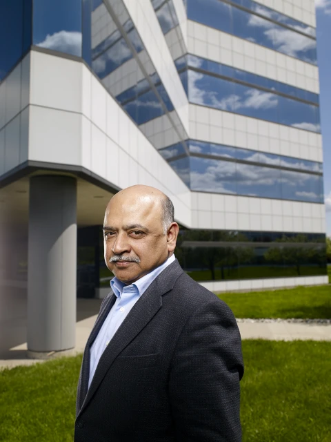 Portrait of a middle-aged man in a suit and button-down shirt standing in front of an office building and a green lawn.