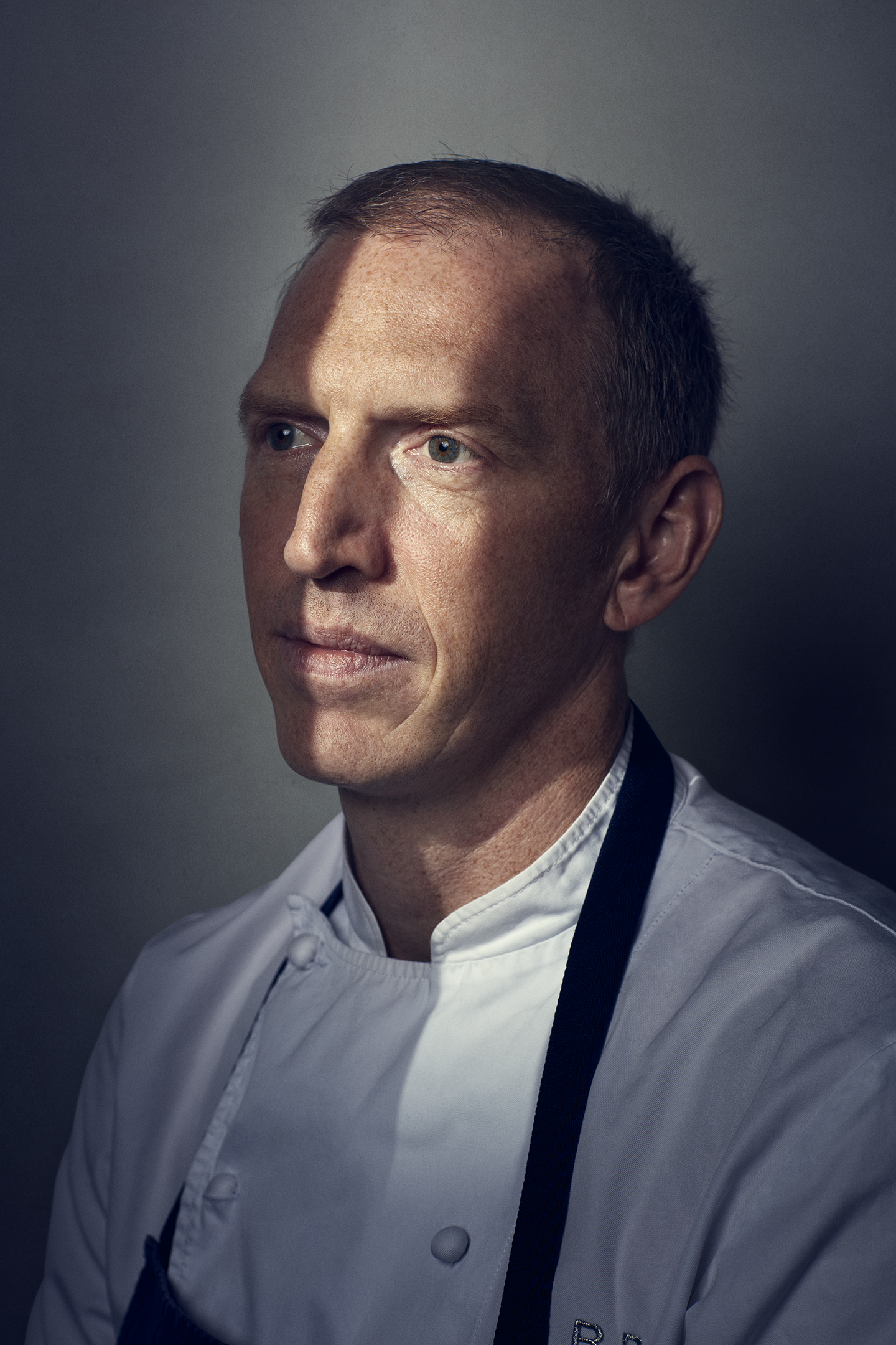 Portrait of a man in a chef's uniform with a dramatic lighting effect, highlighting his face against a simple, grey backdrop.