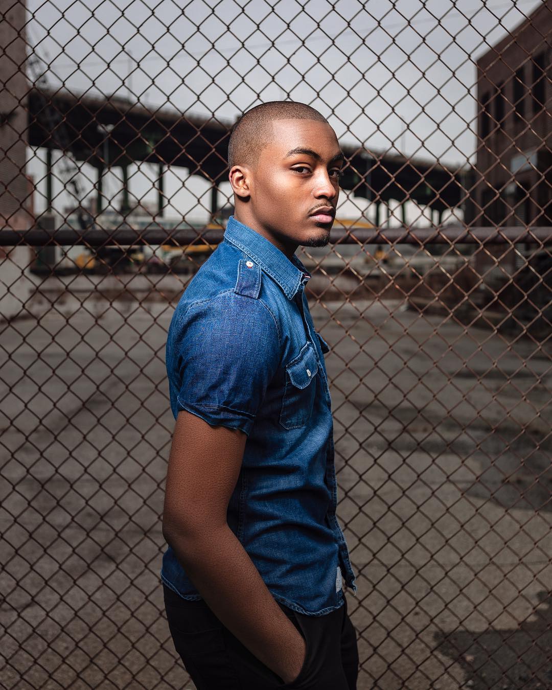 Portrait of a dark-skinned man in a blue denim shirt standing in front of a wire fence, with brick buildings in the background.