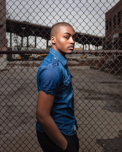 Portrait of a dark-skinned man in a blue denim shirt standing in front of a wire fence, with brick buildings in the background.