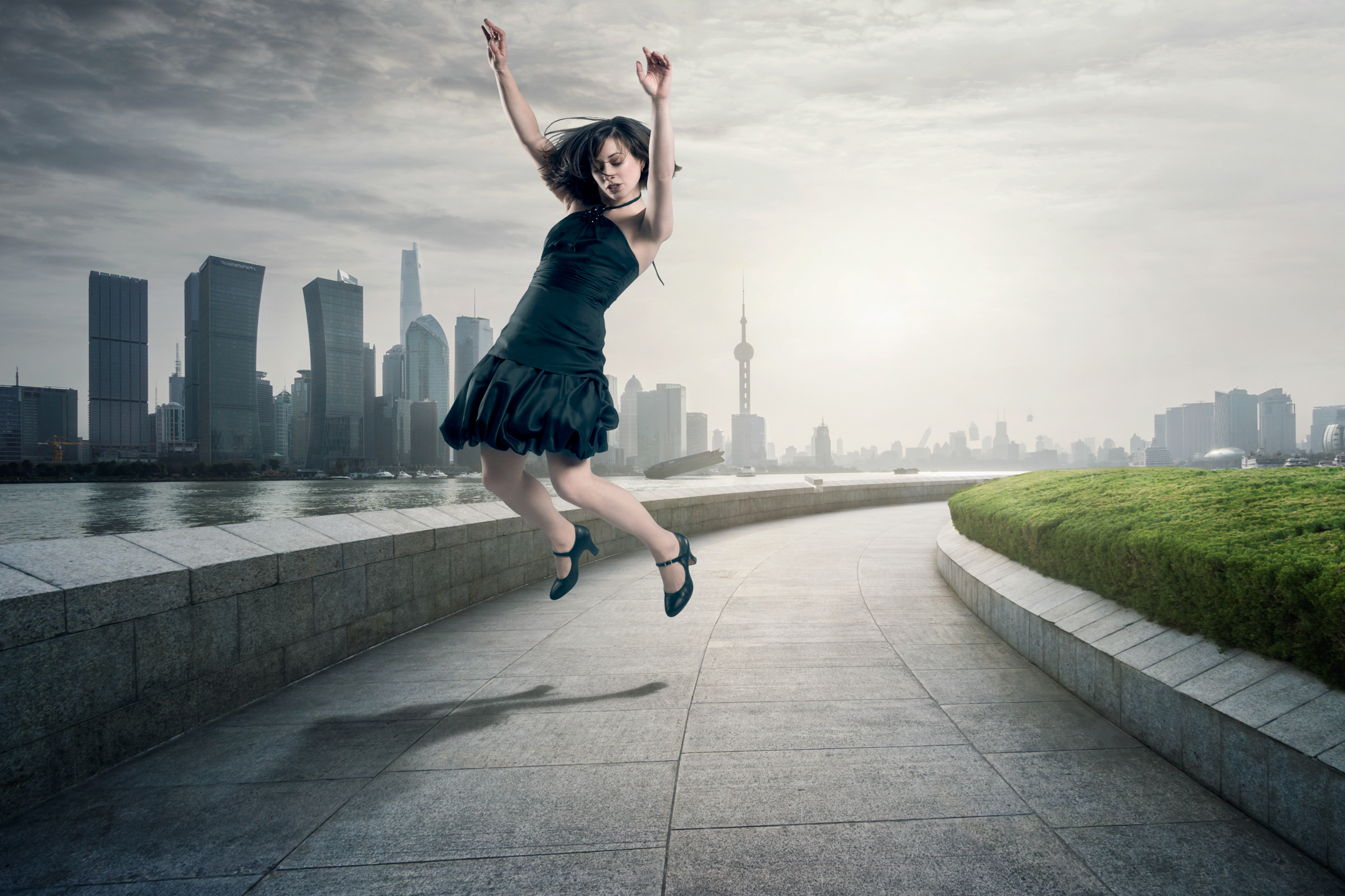 Woman in a black dress leaps in front of a modern city skyline on a cloudy day, captured in a dynamic and fashionable pose.