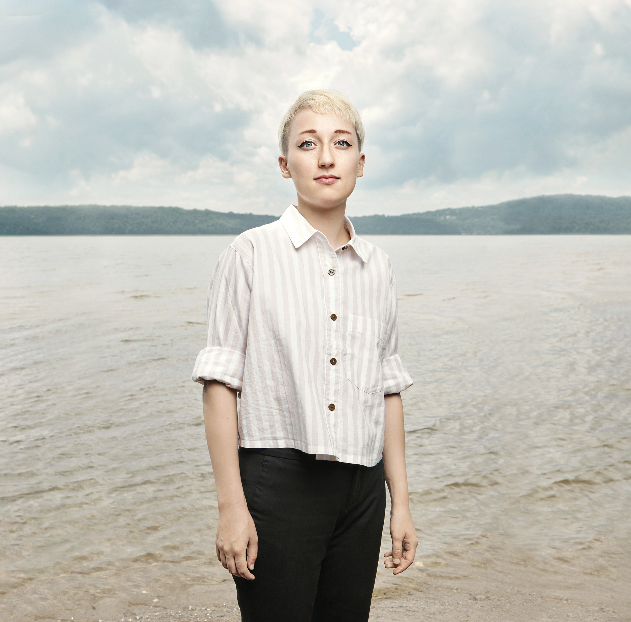 Eye-level portrait of an adult female with short blonde hair, wearing a button-down shirt, standing in front of a lake and cloudy sky.