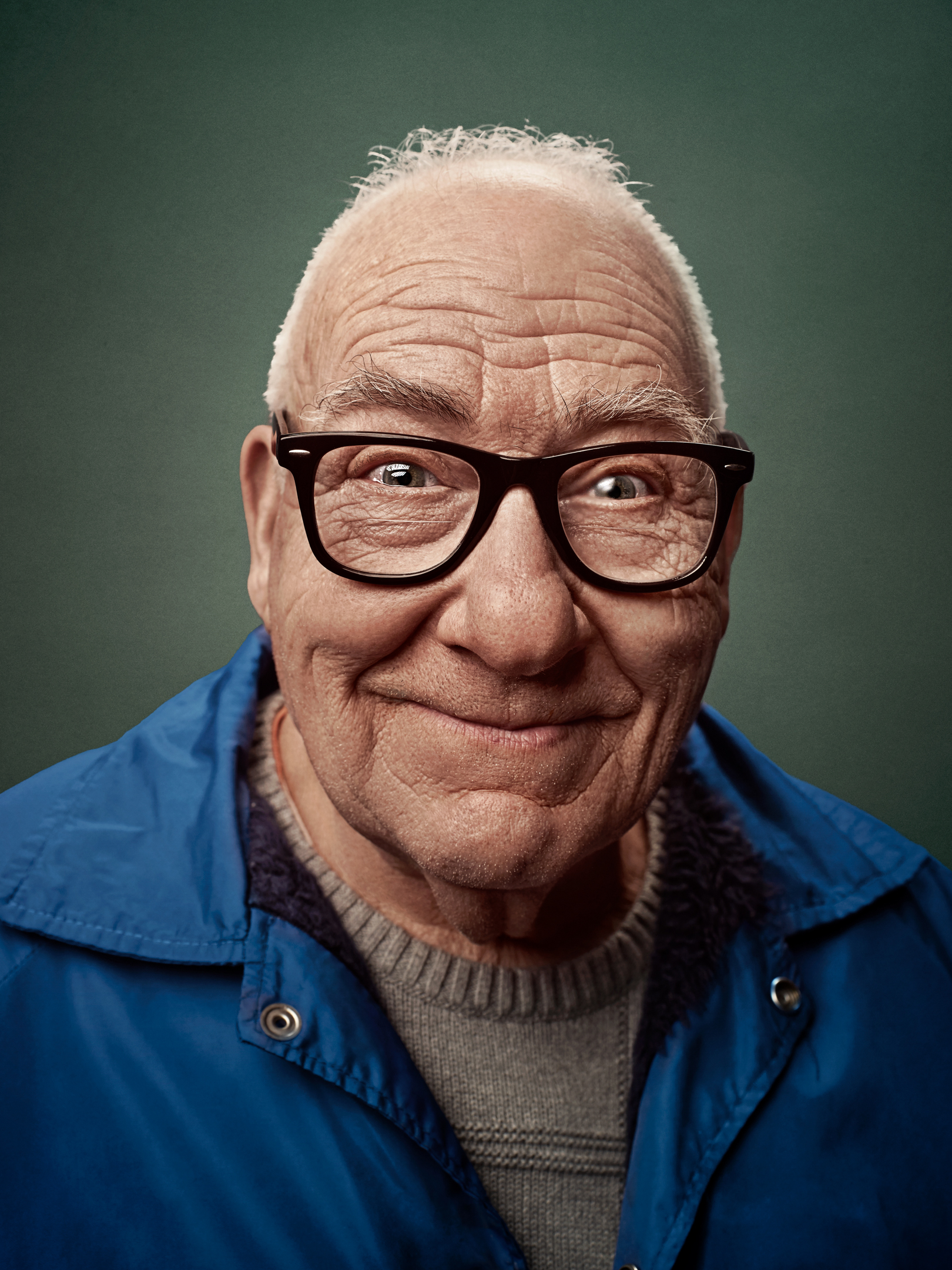 Portrait of an elderly man with a smile, wearing black glasses and a blue jacket, set against a solid green backdrop in the studio.