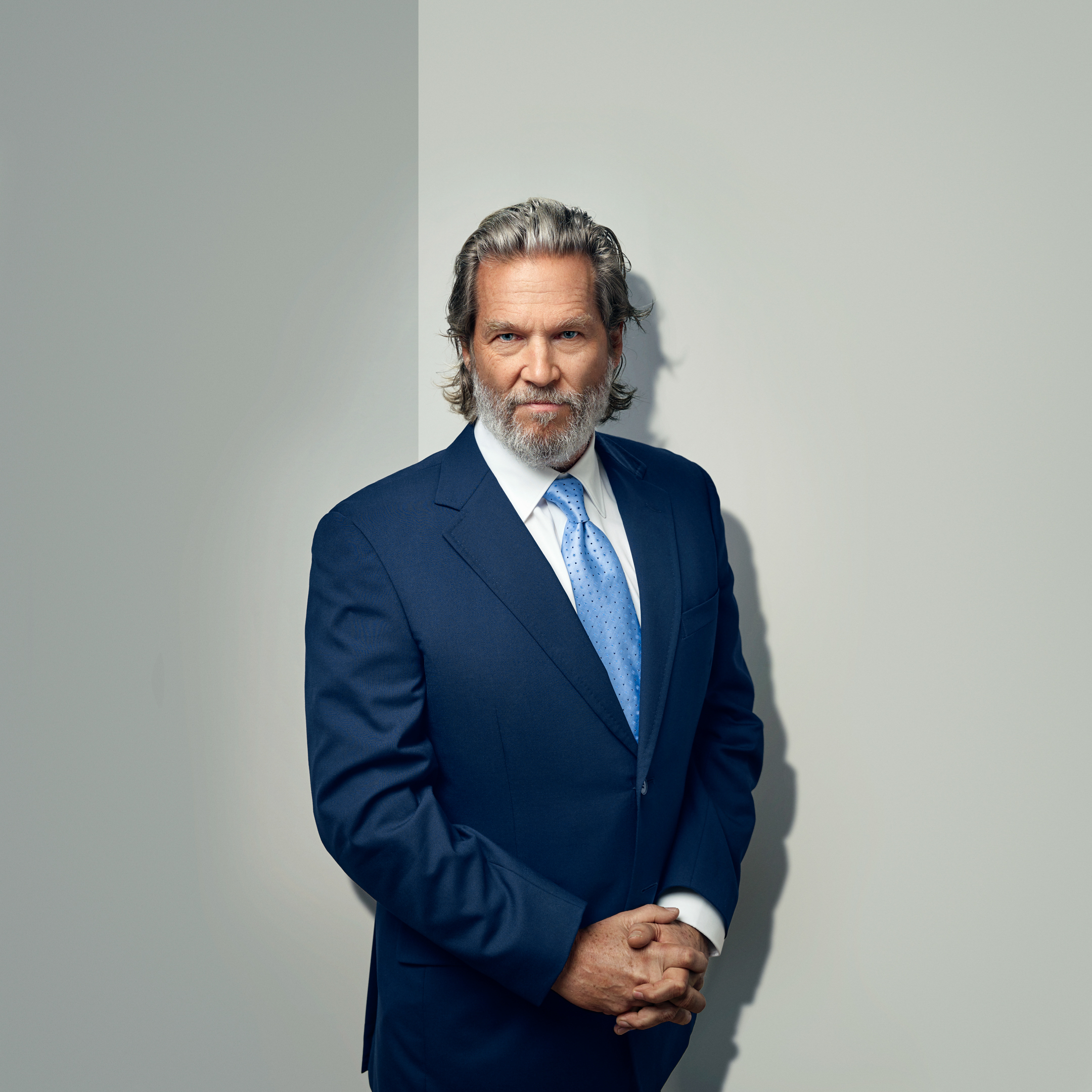 Portrait of older man in blue suit with light blue tie, grey hair and beard, standing indoors in front of a plain wall.