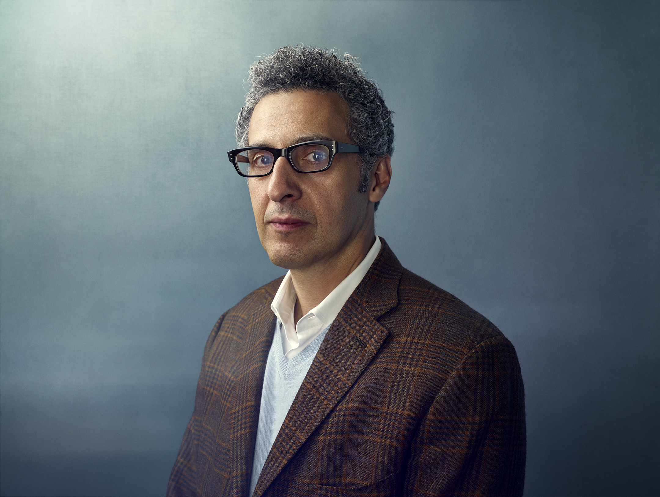 Portrait of an adult male with grey curly hair and glasses wearing a brown plaid blazer against a gradient blue background.